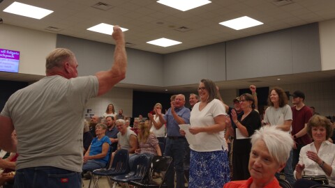 A man in a grey shirt faces a crowd of clapping and standing people. He has his fist in the air as a sign of victory and other people pump their fists as well. The people are in a school cafeteria. 