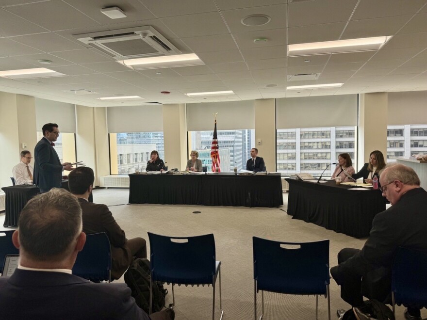 A man stands and speaks to a panel of five people seated at tables in a conference room, with an American flag in the background and several attendees seated facing the panel.
