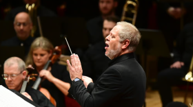 Man in suit with grey hair conducts group of people holding instruments 