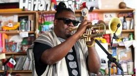 A man that has his locks in a bun while wearing sunglasses blows into a trumpet. 