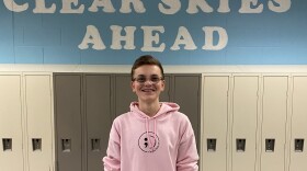 A high schooler in a pink sweatshirt and with braces and glasses smiles at the camera with his hands in his pockets. He stands in front of a bay of lockers with a baby blue mural above that reads "Clear Skies Ahead"