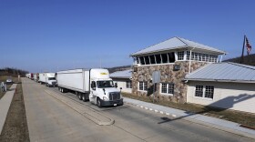 Trucks line up to pass through a weigh station. The weigh station looks like a driveway with a stone building. 