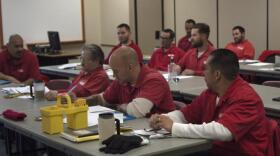 Men in red prison clothing at classroom desks listen to a speaker.