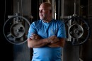 A man in a blue t-shirt stands in front of industrial-looking equipment with his arms crossed. Grain Merchandiser Scott Harre at the Top AG Grain Co-Op in Okawville, Ill. Harre started working at the grain elevator more than 30 years ago after receiving a degree in accounting. 