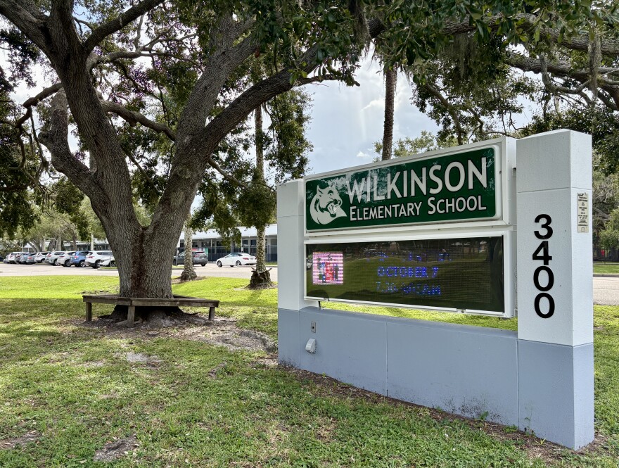 A school sign on grass says Wilkinson Elementary School with a large tree to the left