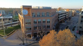 A large four-story brick building with tall windows in an urban setting along a tree-lined road with railroad tracks behind and a large analog clock on the top floor of the building and a sign that reads 'Uptown Station' at the entrance.