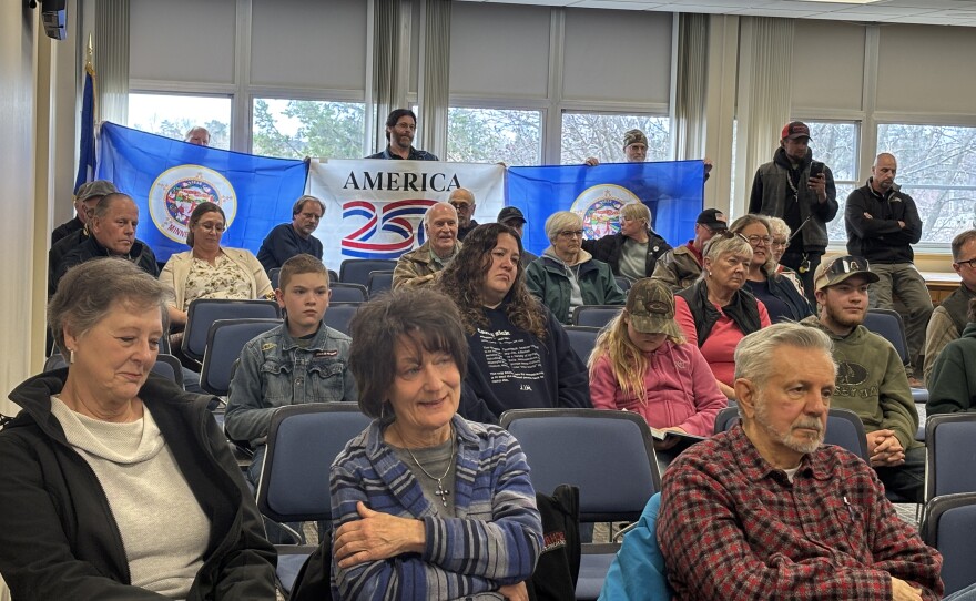Three attendees hold two former Minnesota state flags and an "America 250" flag May 5, 2026, in the back of the Itasca County Boardroom in Grand Rapids.