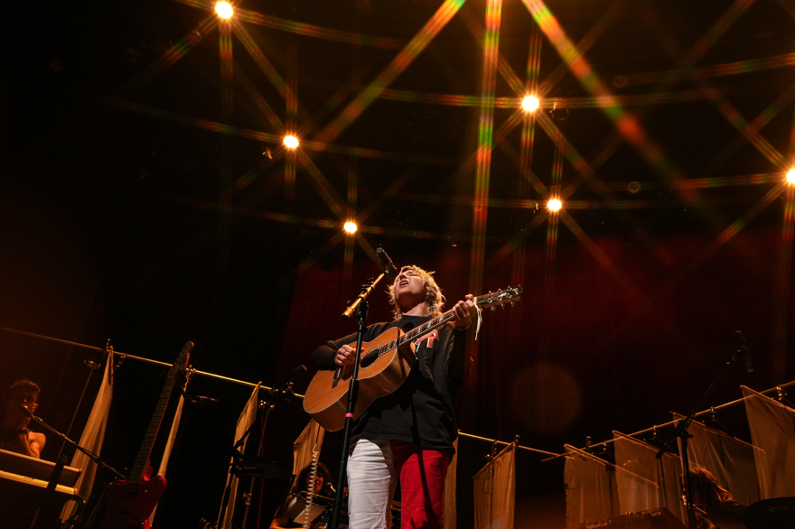 A distant shot of Caroline Rose, looking up at her while she performs on stage. Sheer white curtains are in the background.