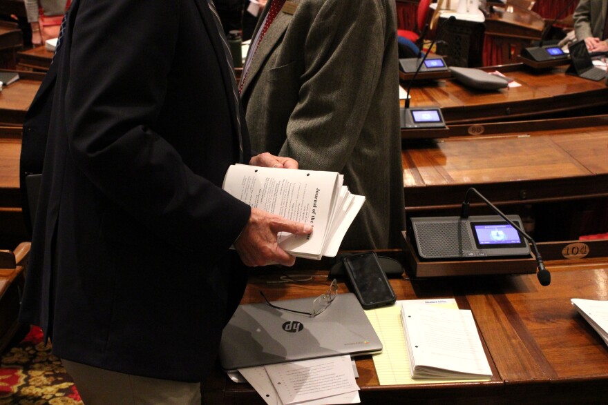 A hand holding a stack of papers. Glasses, a computer and more papers sit on a desk behind.