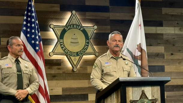 A man in a sheriff's uniform stands behind a podium next to another man in the same uniform. 
