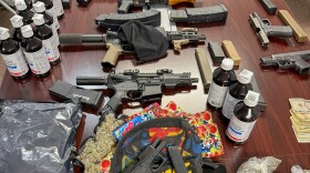  Multiple firearms, magazines, bottles, bags of plant material and cash are arranged across a table.