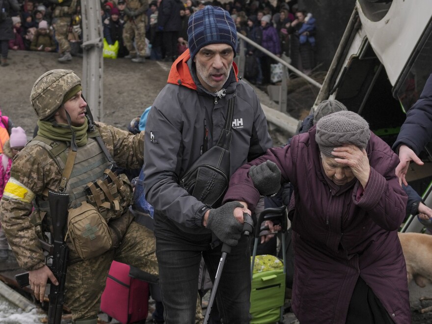 An elderly woman is assisted while crossing the Irpin river, under a bridge that was destroyed by a Russian airstrike, as civilians flee the town of Irpin, Ukraine, Saturday, March 5, 2022.