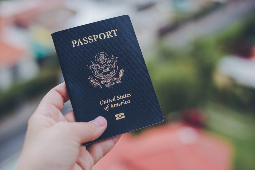 A hand holding up a dark blue United States passport, with a blurred street scene of buildings in the background.