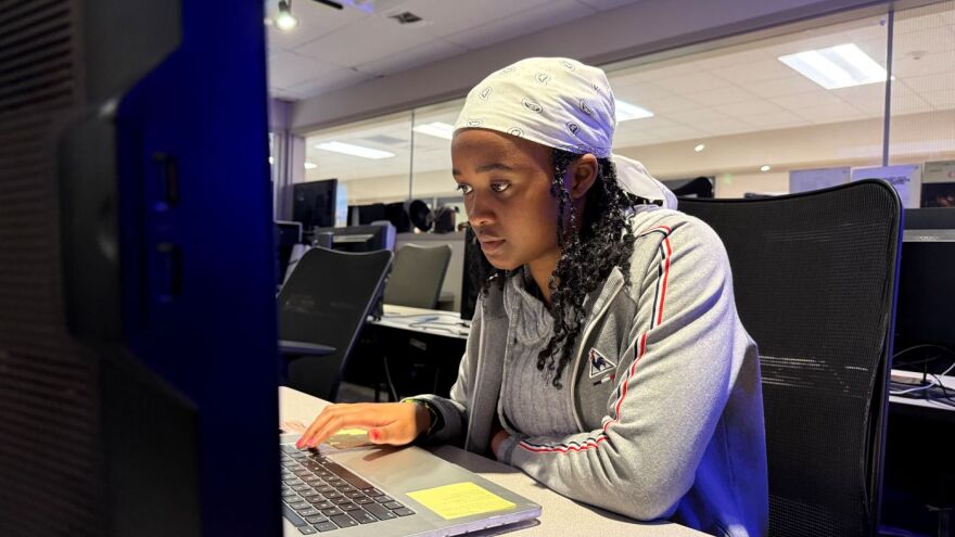 A young woman wearing a white bandana over her black braided hair is looking at a computer.