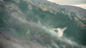 A yellow and red super scooper plane, dumping water over smoke emerging from mountainous terrain