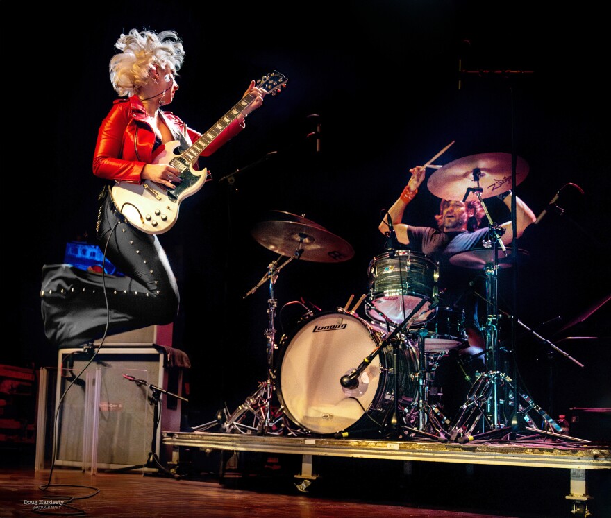 Female guitarist leaping the the air in front of the drummer.