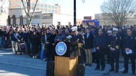 Buffalo Firefighter Commissioner William Renaldo stands outside behind a podium with the Seal of Buffalo on it. He wears a black jacket with gold symbols on it and a white cap. Firefighters and the family of Jason Arno stand behind him on the sidewalk.