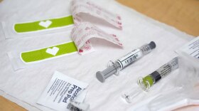Medical supplies including syringes, vials, adhesive bandages, and alcohol pads are arranged on a table, presumably for a vaccination or medical procedure.