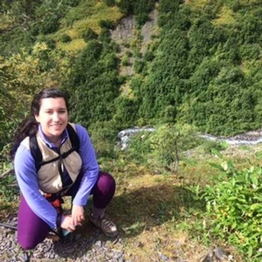 A woman wearing a backpack and purple clothing kneels for a photo in front of a wooded area
