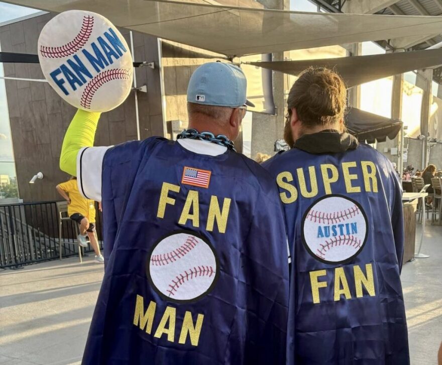 Two men stand next to each other wearing capes. The one on the left says "Fan Man" while the one on the right says "Super Fan." 