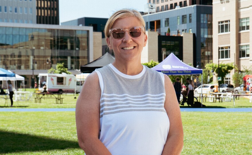 A woman with blonde hair wearing sunglasses, a grey and white striped tank top, and black pants. She is standing in front of a green space with buildings in the background. 