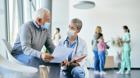 Mature patient communicating with female doctor about medical examination data at the clinic and wearing protective face masks due to COVID-19 pandemic.