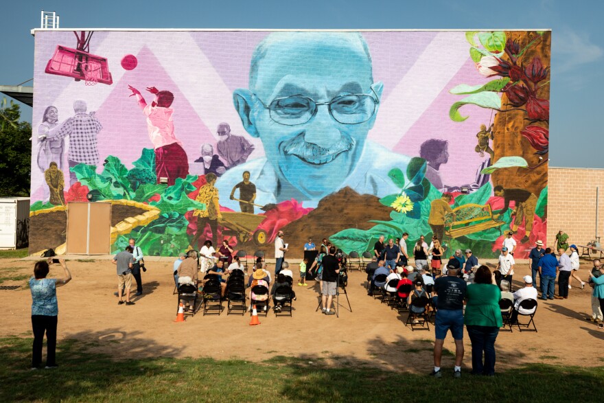 People sit in rows of chairs in front of a blue and pink mural of Gus Garcia.