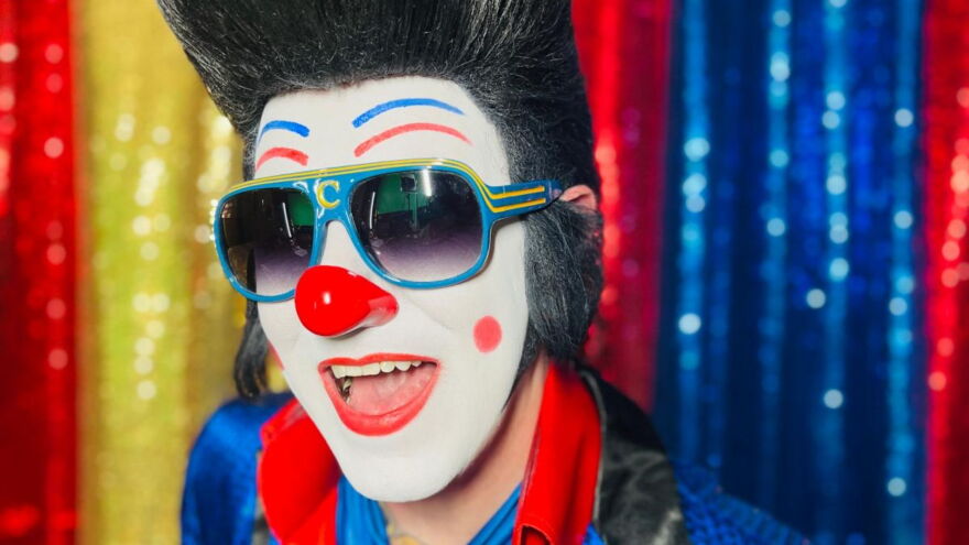 A man with clown face make up also looks like Elvis Presely with a fake black updo wig and a red nose. He is smiling.
