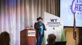 A man wearing a cowboy hat and a black blazer speaks into a microphone as he stands next to a podium and a sign that reads “WY Governor’s Hospitality & Tourism Convention.”