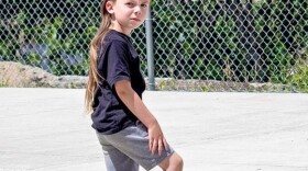 A little boy with long light brown hair, a black shirt and grey shorts stands on a black skateboard. Behind him is a chain link fence.