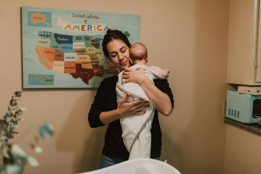 A woman holds a newborn wrapped in a blanket against her chest. There's art of the U.S. states on the wall behind her, stylized to look more like something from a children's picture book.