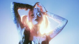 A woman with tattoos on her arms and artist makeup on her face stands with her hands on her head with a spotlight directly behind her.