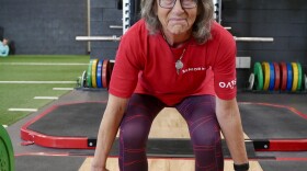 An older woman in a red t-shirt, purple workout leggings and blue grippy shoes demonstrates how to do a deadlift, all with a smile on her face.
