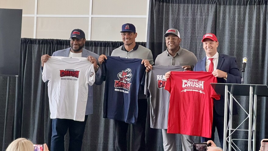 Four men standing on a stage holding up t-shirts with a grey curtain behind them.