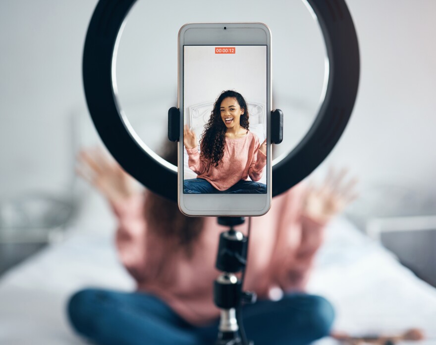 Live streaming, phone and black woman talking on video podcast in the bedroom of her house. Happy and excited girl or influencer speaking on the internet or social media with a mobile and ring light.