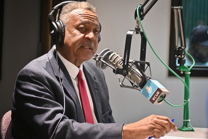 A man wearing a gray suit and red tie sits inside a radio studio. He is talking at a microphone with a "KCUR" flag on it.