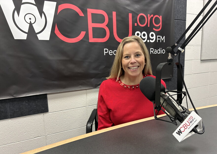 A woman in a red sweater sits in a radio studio in front of a microphone, smiling. Behind her is a WCBU 89.9 FM banner on the wall.