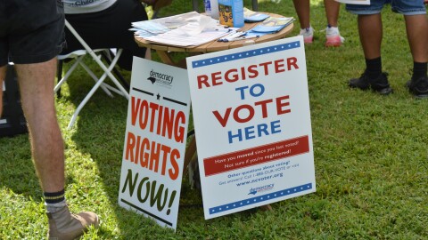 Two signs read "Democracy, voting rights now!" and "Register to vote here."
