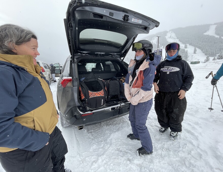 A woman with short gray hair stands on the far left in a snow covered parking lot wearing a blue and gold ski jacket. In the background is Copper Mountain with two groomed trails. In the center of the photo is the back of an SUV with the rear door open and three ski boots bags sit in the back. A teenage girl wearing ski boots, blue ski pants and a helmet with goggles, zips up her blue, light pink and rose pink ski jacket. Next to her stands a teenage boy wearing ski boots, black ski pants, a black sweatshirt with a white "Boulder" logo, blue neck and ear warmer, helmet and googles. On the far right is an arm and had carrying two ski poles.