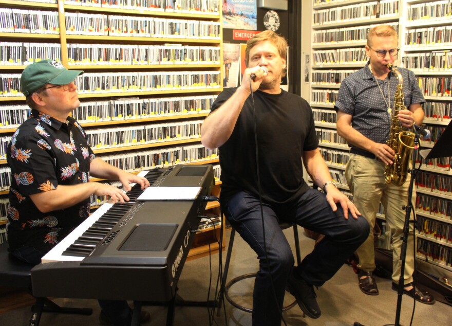 A man plays keyboard to the left of a man sitting on a stool singing into a microphone. On the right is a man playing saxophone.