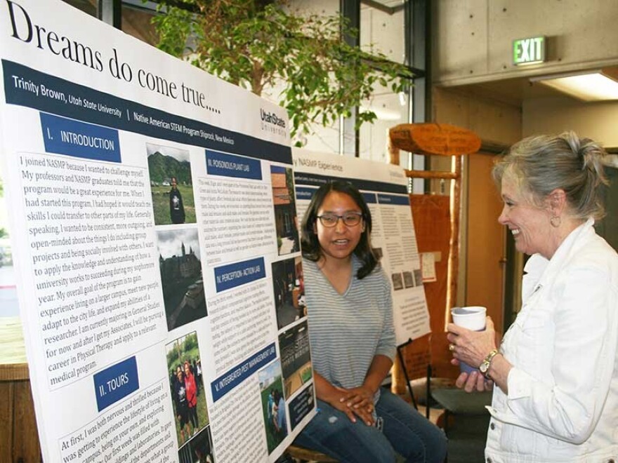 A Native American scholar presents her research to the Science college dean at Utah State University. White poster with photos and descriptive text about the student's research.