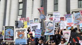 A group of people hold colorful protest signs in front of a light grey building with columns. A couple signs say "WATER IS LIFE DEFEND GREAT SALT LAKE" AND "UPSTREAM & DOWNWARD."