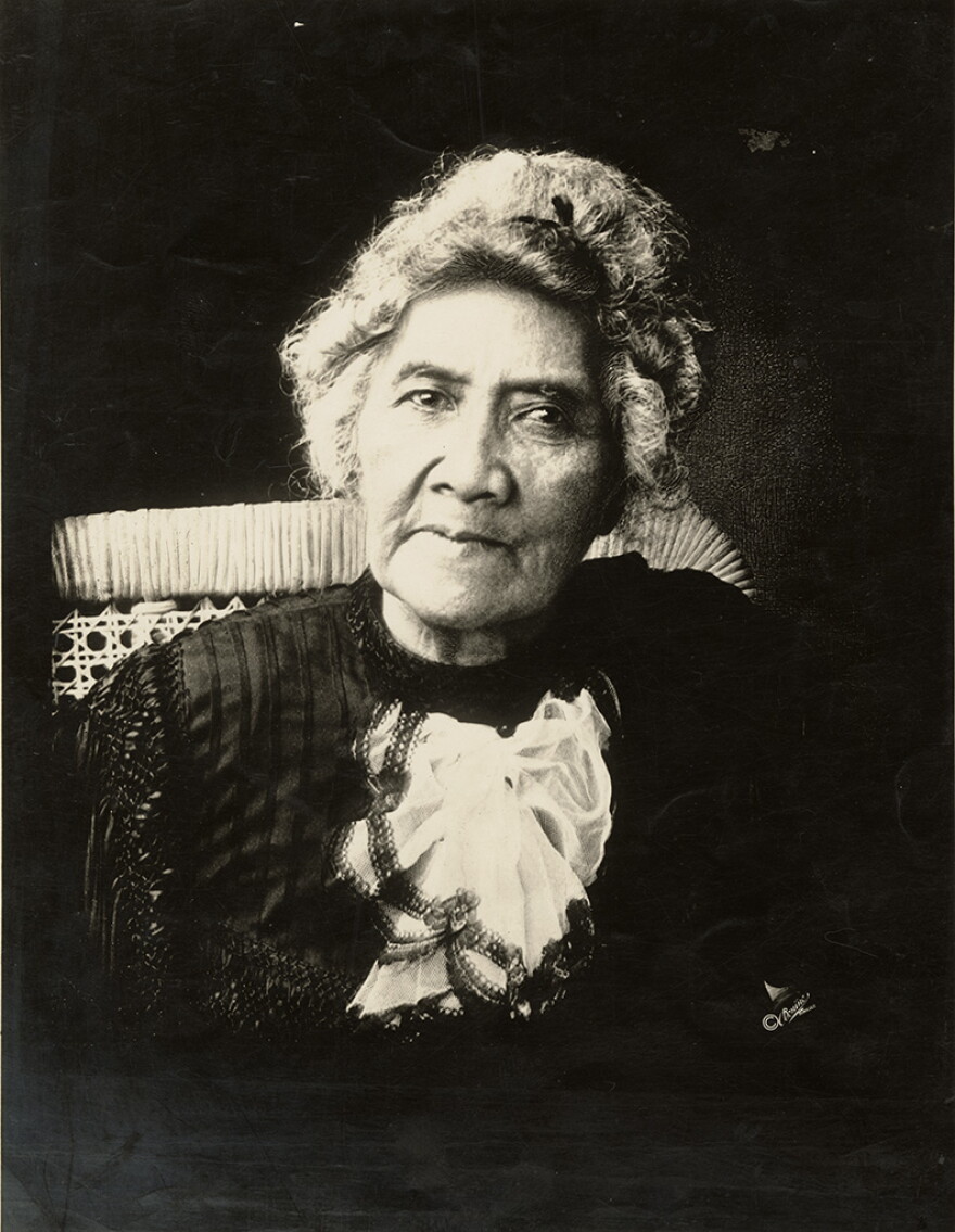A black-and-white photograph of Queen Lili'uokalani in her older years. Her gray hair is tied up in a ribbon and she sits in a wicker chair. 