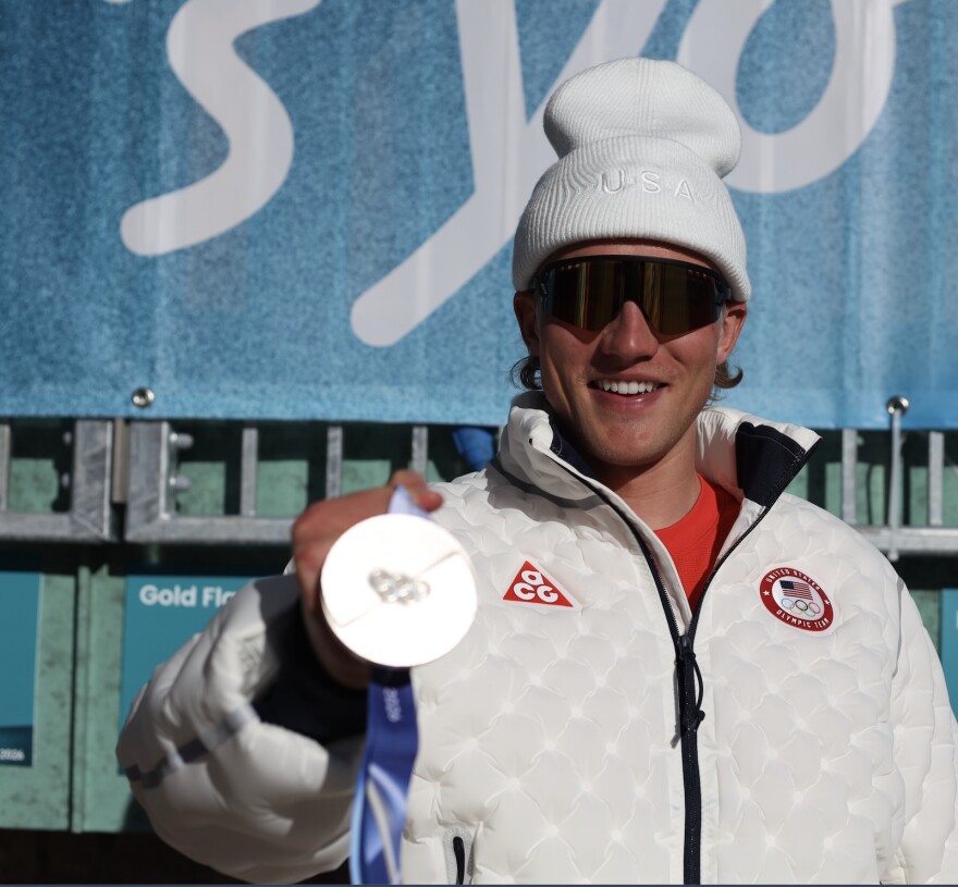 A man in white winter clothes and sunglasses holds up a silver medal.