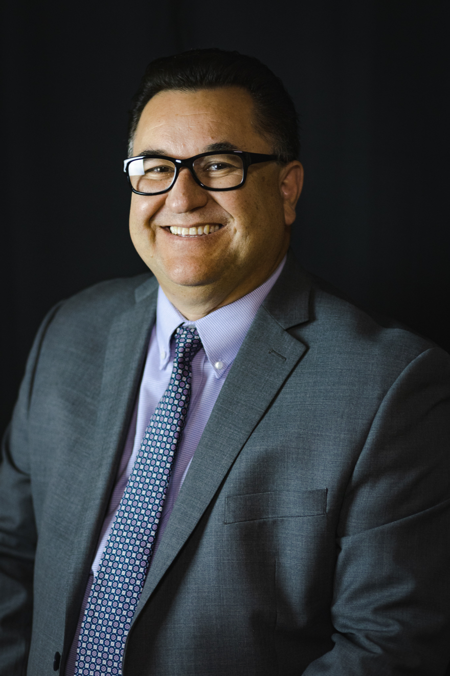 A portrait of a smiling man, with a black background. He is smiling, wearing a blue suit and tie, and dark rimmed glasses. 