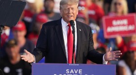a man in a suit, white shirt and red tie speaks at a podium with a crowd behind him