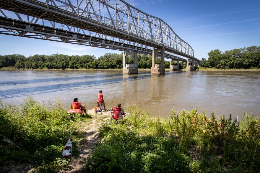 Three people in red shirts fish off the leafy banks by a river, under a bridge. 