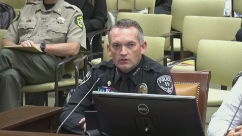 A police officer sits behind a microphone. The room behind is filled with chairs. 