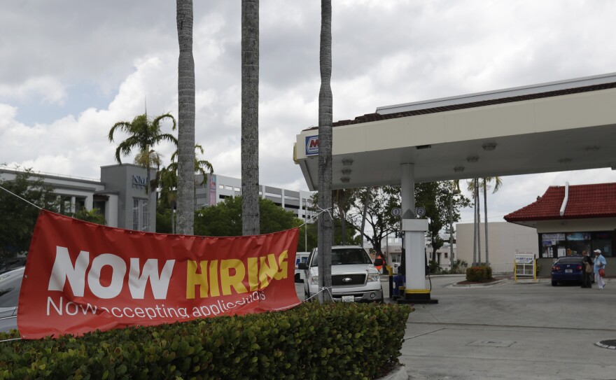 A big banner is tied up on trees. It reads, "Now hiring. Now accepting applications." Besides the banner is what looks to be a gas station.