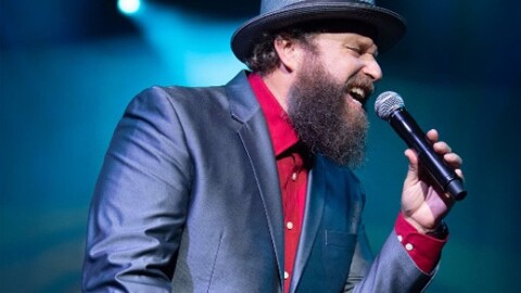 A bearded male singer in a gray suit, red shirt, and gray hat performs energetically on stage.
